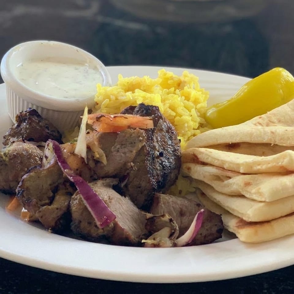 Plate of grilled meat with yellow rice, stacked flatbreads, pickled pepper, sliced onions, and creamy dipping sauce.
