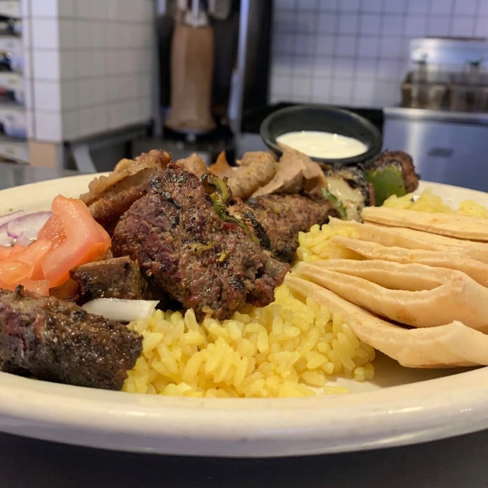 Mediterranean mixed grill platter with skewered meats, yellow rice, pita bread, salad, and dipping sauce in a restaurant kitchen.